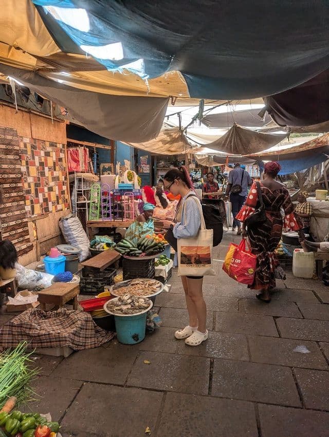 Una donna con una borsa tote si aggira in un corridoio affollato di un mercato coperto, osservando bancarelle di prodotti freschi e frutti di mare.