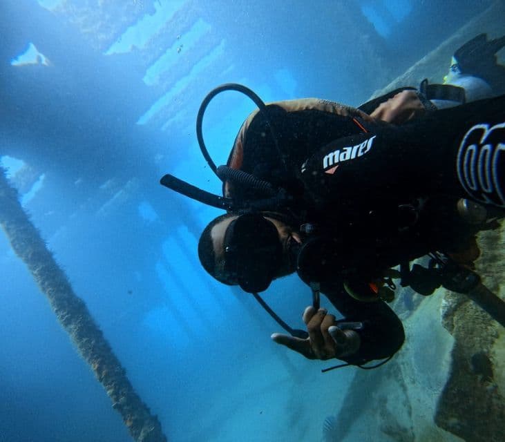 Un subacqueo con muta nera nuota sott'acqua accanto a una struttura sommersa in acque limpide e blu.