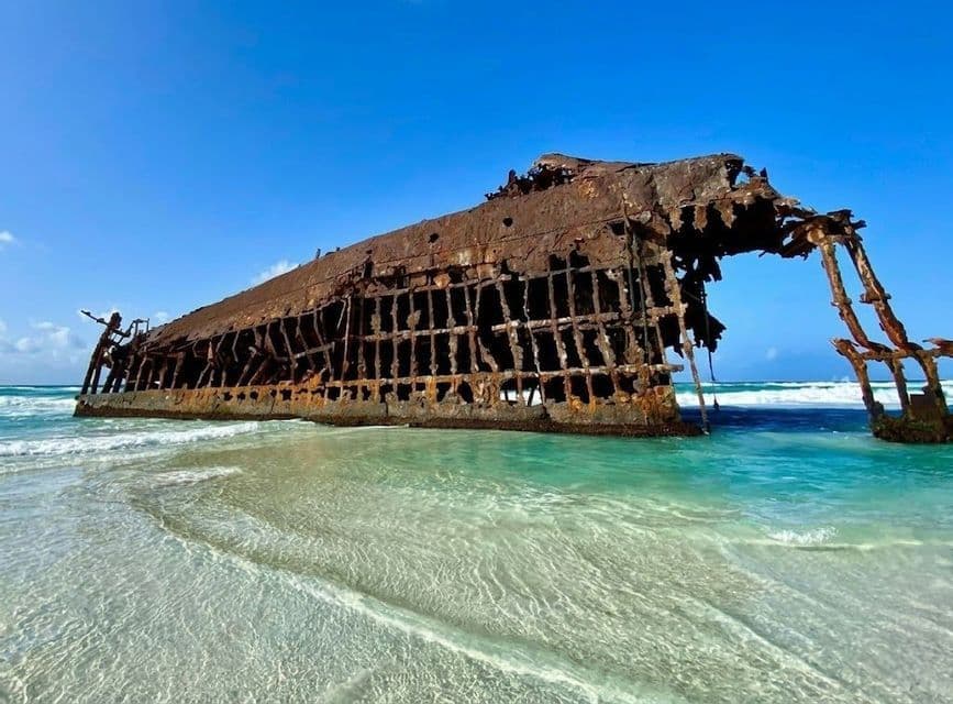 Lo scafo arrugginito di un grande relitto giace su una spiaggia sabbiosa, con acqua turchese e limpida che lambisce la riva sotto un cielo blu.