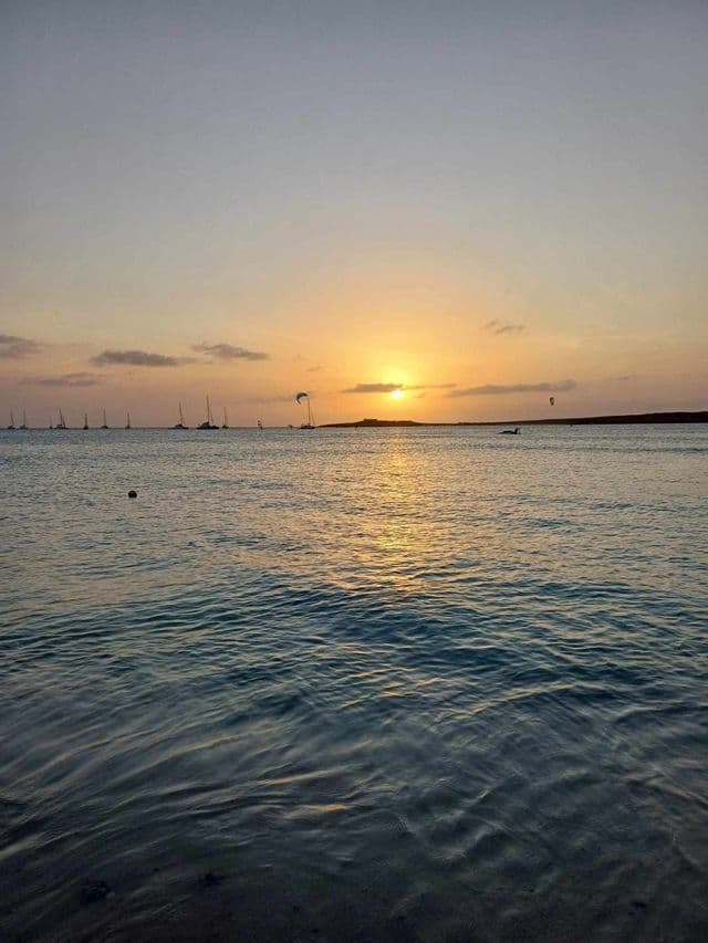 Un tramonto dorato sul mare calmo, con barche a vela e kitesurfer profilati all'orizzonte.