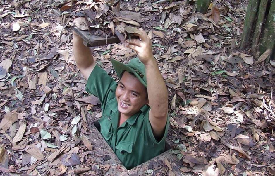 Un uomo sorridente, in uniforme verde e cappello, solleva una botola ricoperta di foglie mentre emerge da un tunnel nascosto nel terreno.