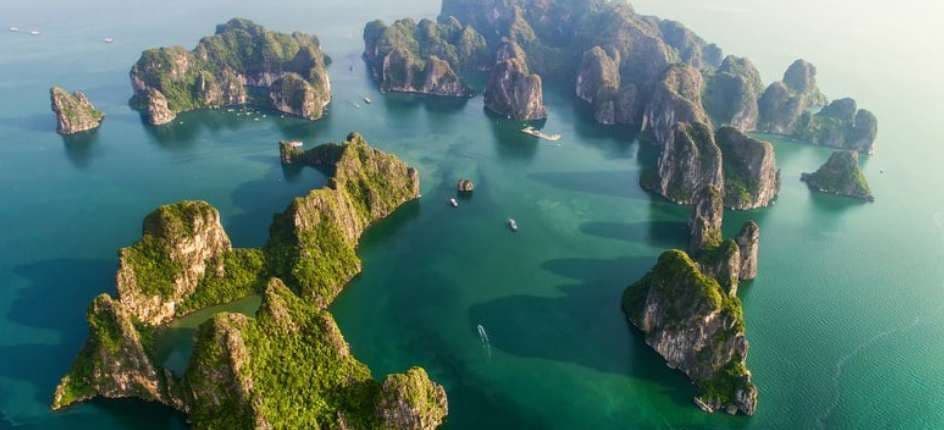Una vista aerea di numerosi faraglioni carsici ricoperti di verde fogliame che emergono da acque calme e verde smeraldo, con piccole imbarcazioni che navigano.