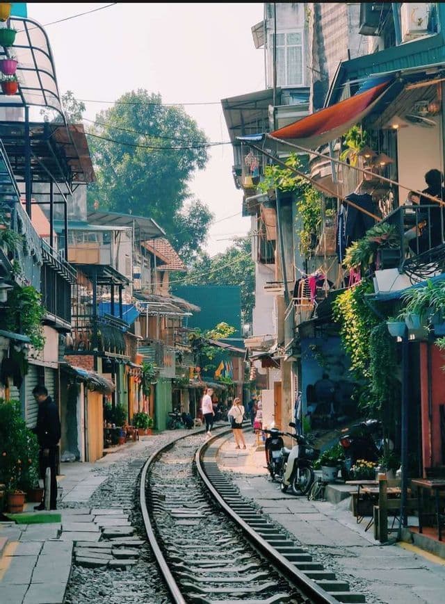 Un binario ferroviario che attraversa il centro di uno stretto vicolo, fiancheggiato da alti edifici colorati con balconi e piante.