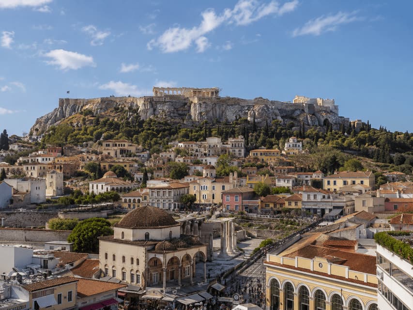 Una vista panoramica di una città storica con antiche rovine su una collina rocciosa che domina edifici e una moschea sotto un cielo azzurro.