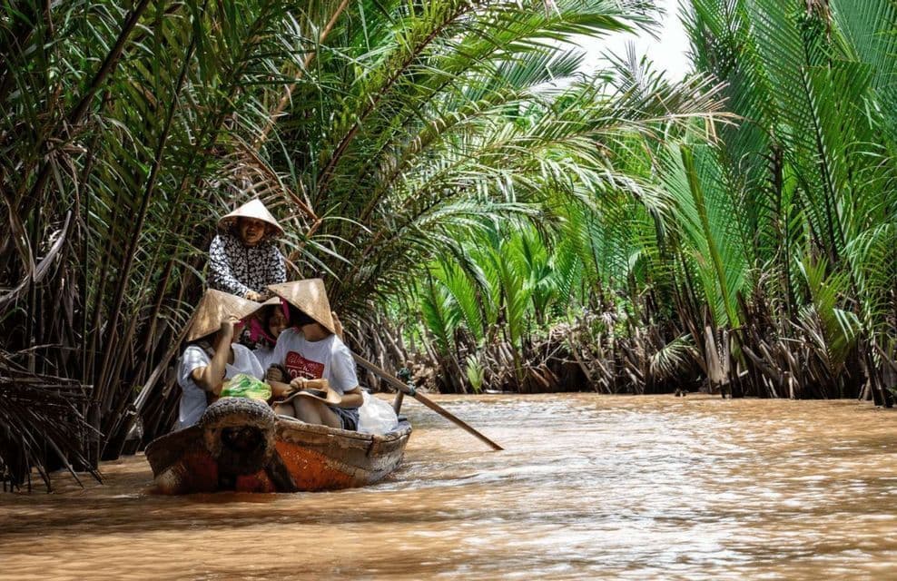Un viaggio di gruppo WeRoad con cappelli conici naviga su una barca di legno lungo un fiume fangoso attraverso una lussureggiante foresta tropicale.