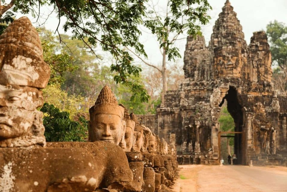 Una fila di antiche statue in pietra con volti scolpiti si allinea lungo un sentiero che conduce a un grande portale di un tempio, immerso nella giungla.