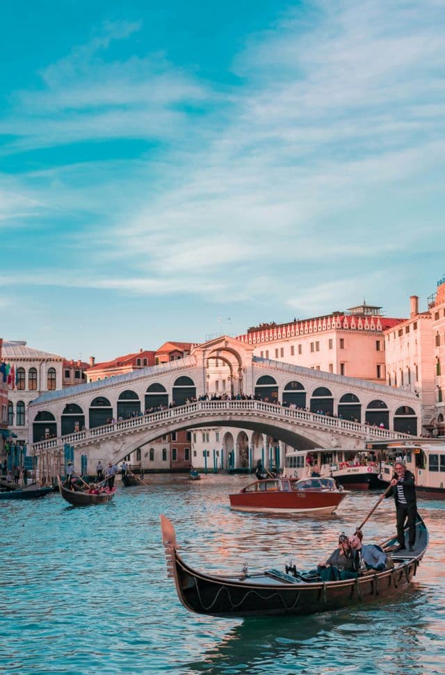A gondolier rows a gondola with passengers on a busy canal, with a large, arched stone bridge in the background under a blue sky.