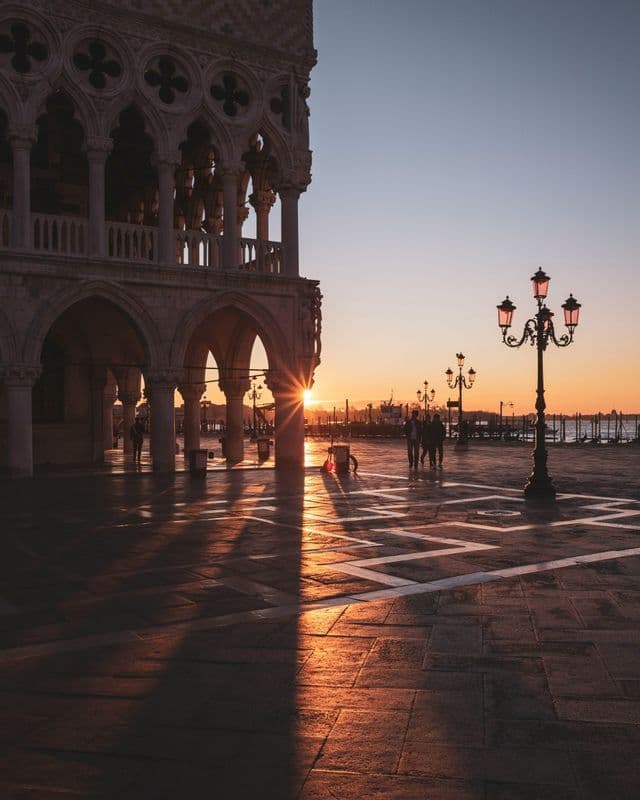 The rising sun creates a starburst through the arches of an ornate building, casting long shadows across a paved plaza with lit lampposts.
