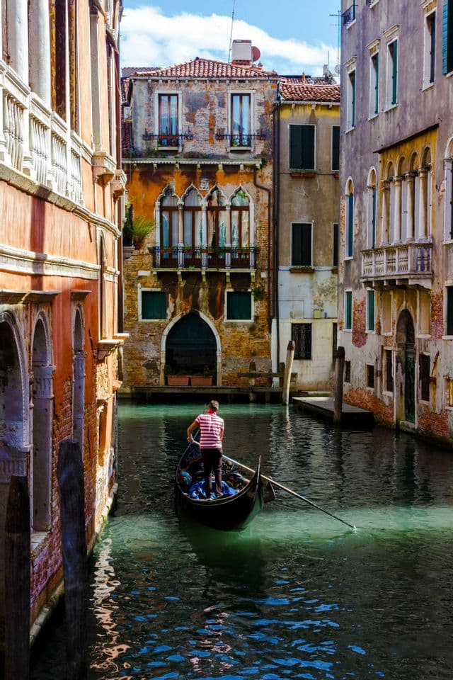 A gondolier in a striped shirt rows a gondola down a narrow canal between tall, historic buildings on a sunny day.
