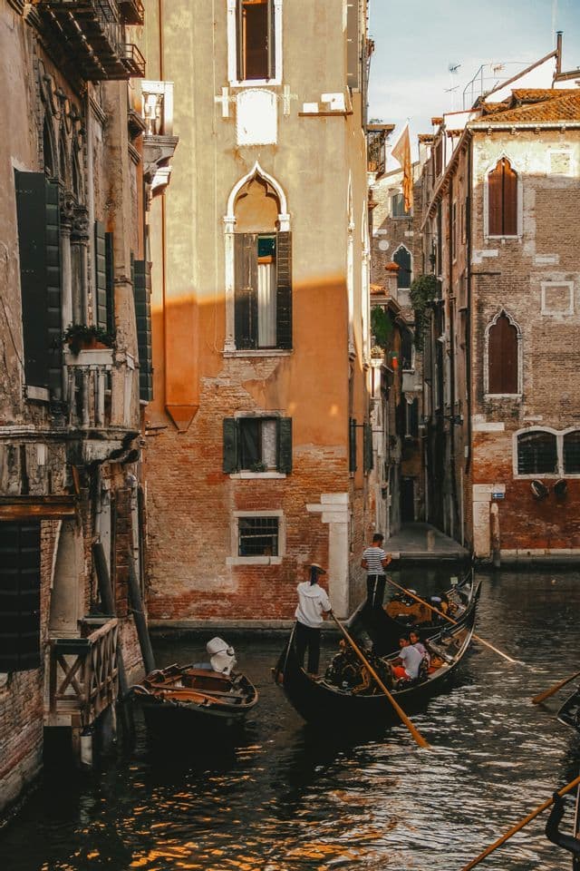 A gondola with a group of people is paddled down a narrow canal between tall, sunlit historic buildings.