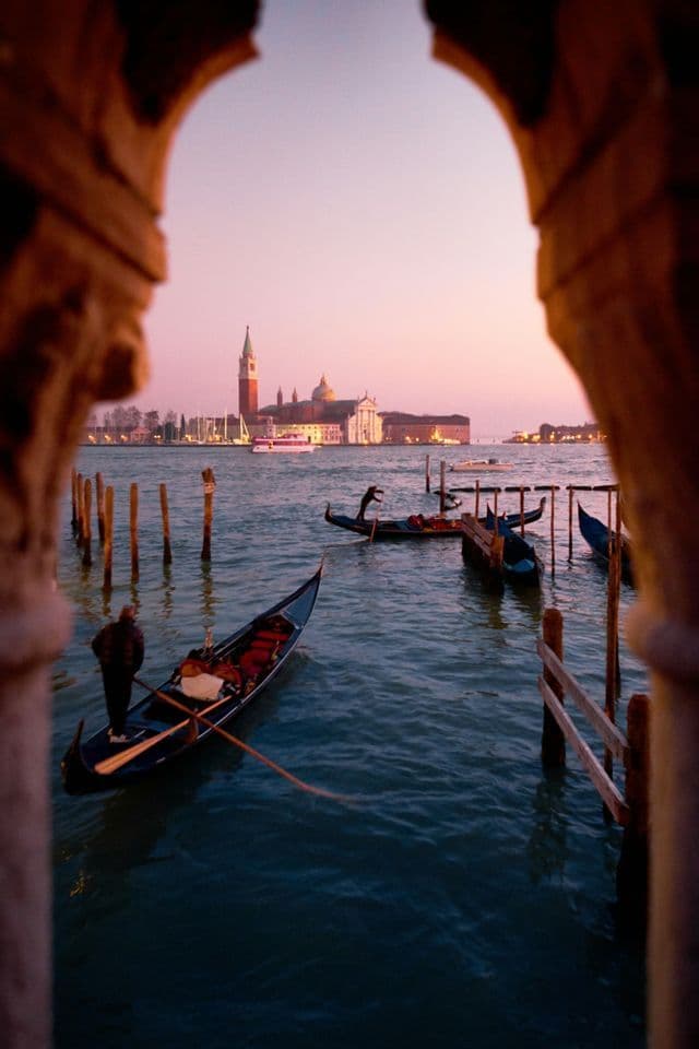 A gondolier rows a gondola on a canal at dusk, with a city skyline visible through a stone archway.