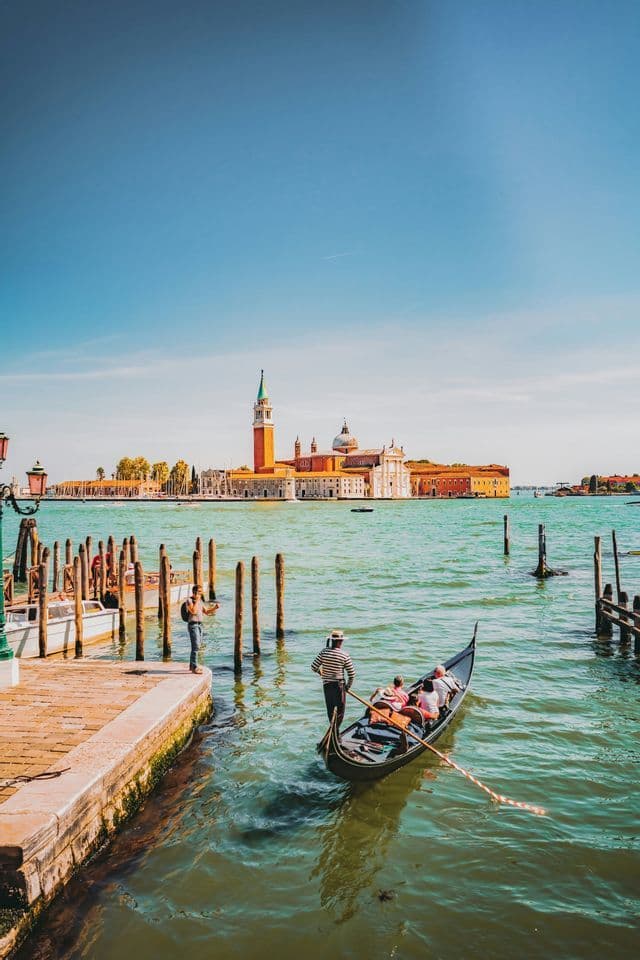 A gondolier steers a gondola with passengers on a canal, with a distant island and bell tower under a clear blue sky.