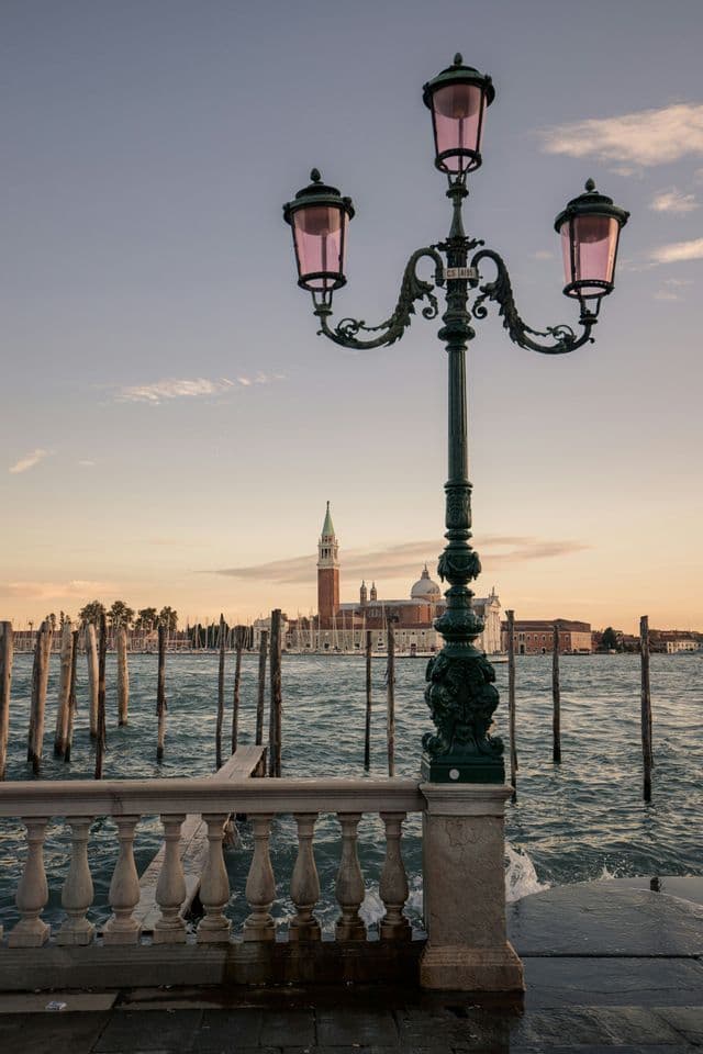 An ornate lamppost with pink glass stands on a stone promenade, overlooking a canal with a bell tower and church at sunset.