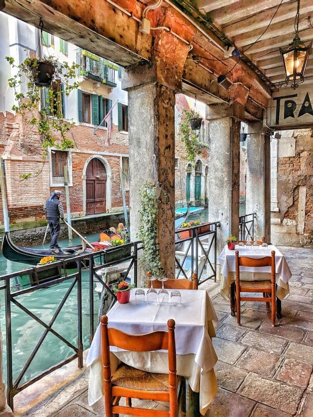 View from a restaurant patio of a gondolier steering a gondola down a narrow canal between historic brick buildings.