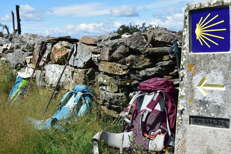Zaini da trekking e bastoncini da trekking riposano nell'erba alta contro un muro di pietra vicino a un segnavia del Cammino di Santiago.