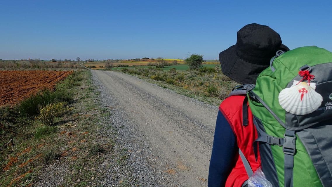 Un escursionista con uno zaino e una conchiglia del Cammino di Santiago cammina su un sentiero sterrato attraverso la campagna.