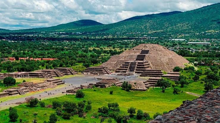 Un'antica piramide di pietra a gradoni si erge in una valle piena di rovine, alberi e verdi colline sotto un cielo nuvoloso.
