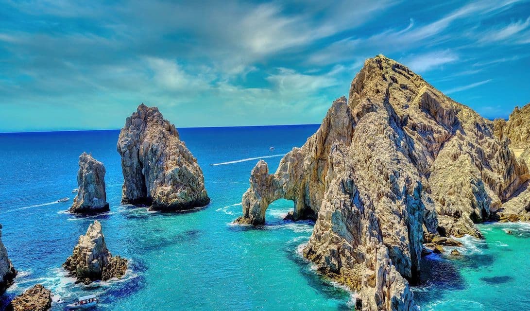 Grandi formazioni rocciose, inclusa una prominente arcata naturale, sorgono da un oceano turchese mentre piccole imbarcazioni navigano sotto un cielo parzialmente nuvoloso.