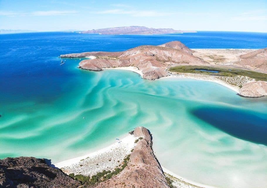 Veduta aerea di una costa rocciosa con una lingua di sabbia bianca circondata da acque turchesi e blu intenso che si increspano.