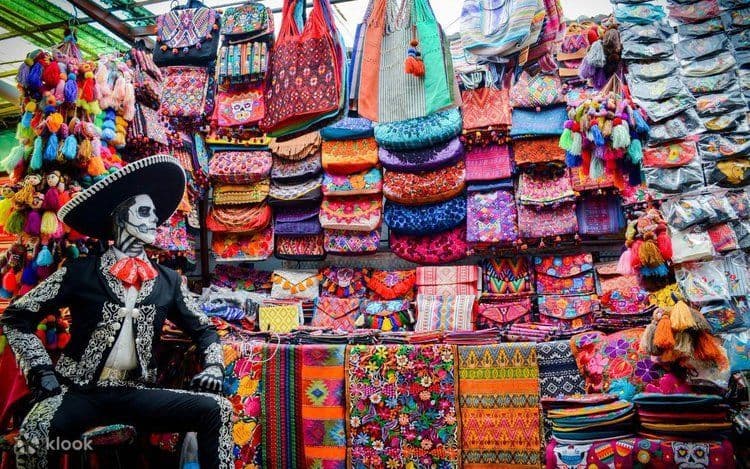 Un manichino in abito da mariachi, con il viso dipinto a teschio, siede a una bancarella del mercato che vende borse e tessuti messicani colorati.