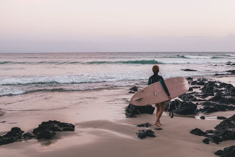 Un surfeur en combinaison de plongée porte une planche de surf rose, marchant sur une plage de sable et de rochers vers l'océan au crépuscule.