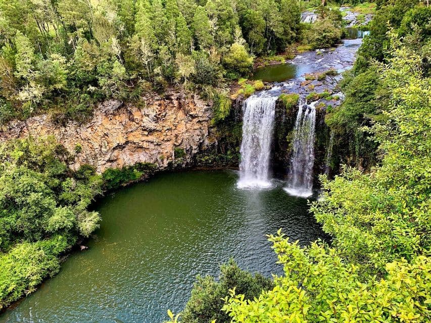 An aerial view of a double waterfall cascading down a rocky cliff into a green pool, surrounded by a dense forest.