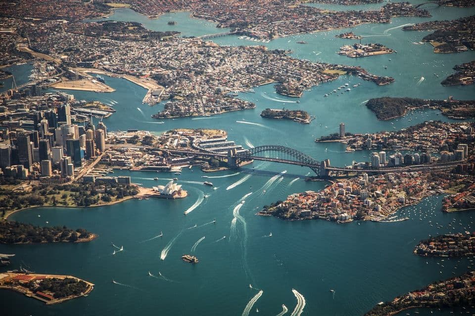 An aerial view of Sydney Harbour, featuring the Opera House and Harbour Bridge, with numerous boats navigating the blue waters.