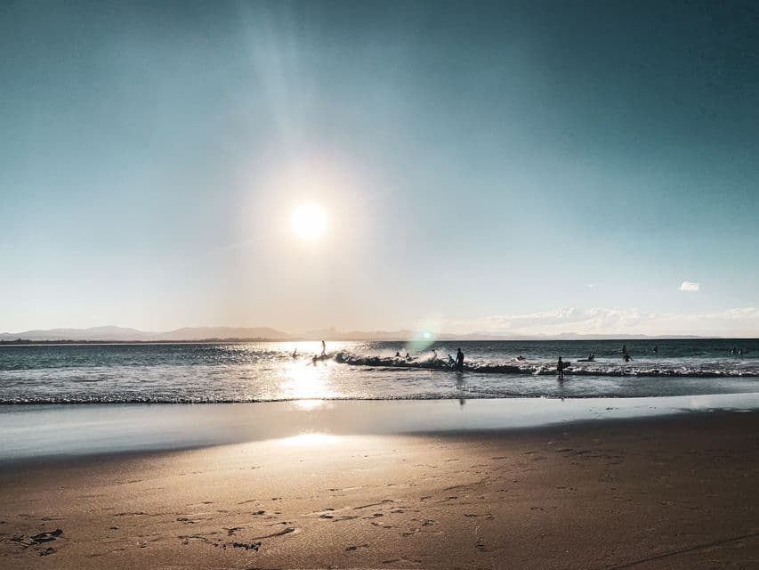 Des surfeurs dans l'océan, chevauchant les vagues au coucher du soleil, vus depuis une plage de sable où le soleil se reflète sur l'eau.