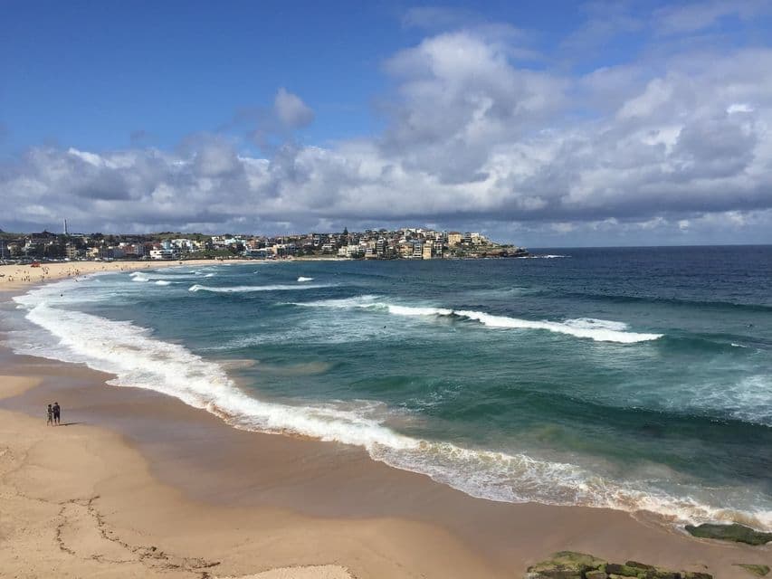 A coastal town overlooks a wide sandy beach as turquoise waves roll onto the shore under a partly cloudy sky.