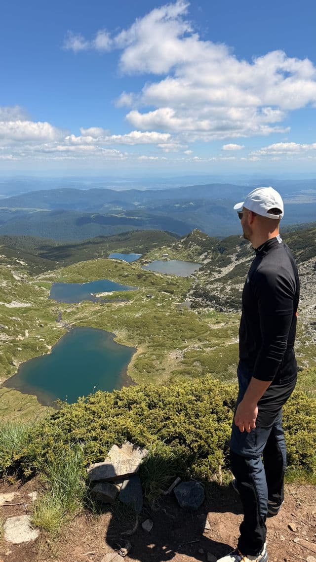Un homme en casquette blanche se tient sur une montagne, contemplant une vallée avec plusieurs lacs bleus.