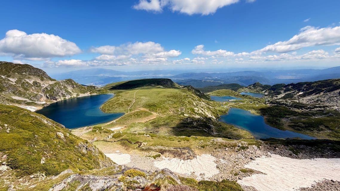 Une vue panoramique sur plusieurs lacs alpins bleus, disséminés dans une vallée de montagne verdoyante, avec des plaques de neige sous un ciel bleu nuageux.