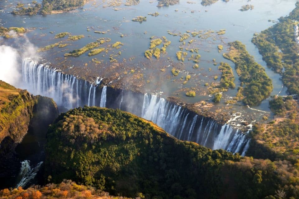 Eine Luftaufnahme eines breiten Wasserfalls, der über eine lange Klippenkante in eine tiefe Schlucht stürzt, umgeben von grünen und gelben Bäumen.