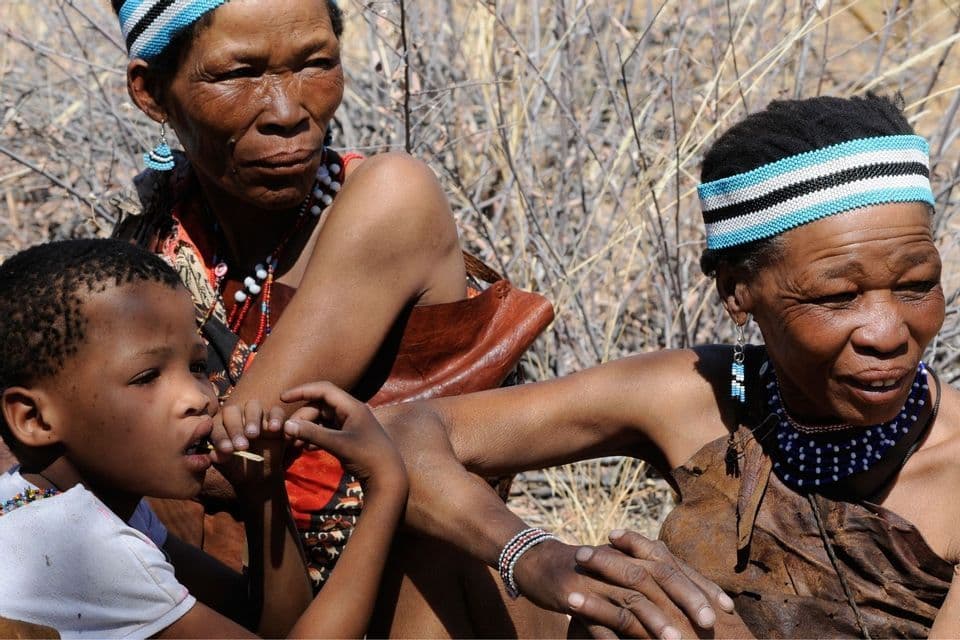 Zwei indigene Frauen und ein kleines Kind in traditioneller Perlenkleidung sitzen zusammen im Freien in einer trockenen, buschigen Landschaft.
