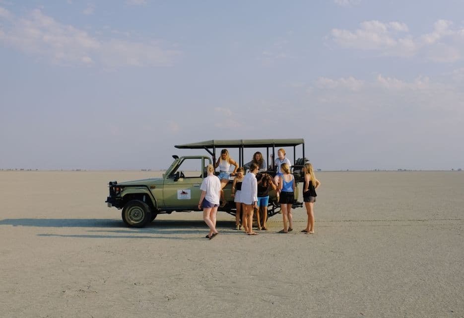 Eine WeRoad-Gruppe steht um einen offenen Safari-Jeep, der auf einer weiten, trockenen Salzpfanne unter einem dunstigen Himmel geparkt ist.