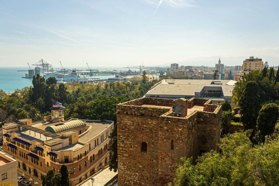 Vista dall'alto di una città costiera con una torre di pietra in primo piano e un porto commerciale con navi sul mare.