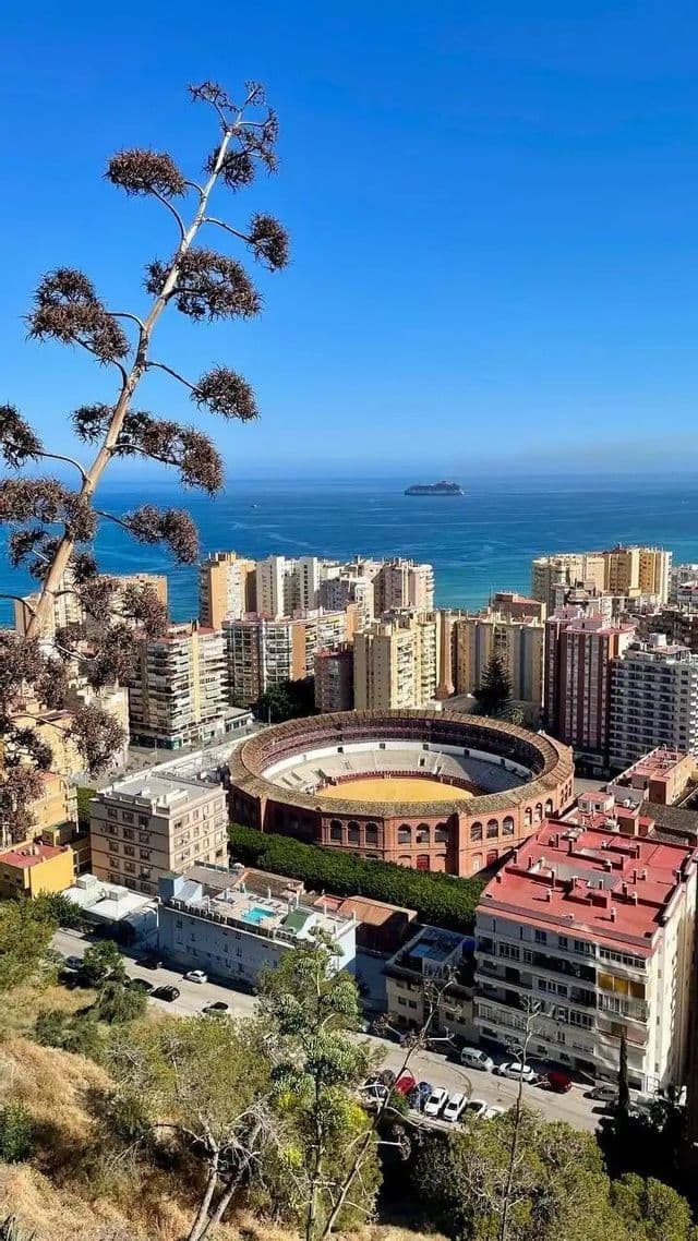 Vue plongeante sur une ville côtière, avec une arène circulaire bien visible, la mer bleue et un navire de croisière à l'horizon.