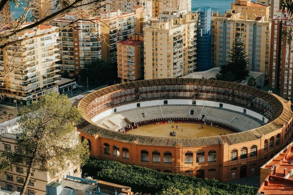 Vue surélevée d'une grande arène circulaire, nichée au milieu de hauts immeubles urbains, avec la mer en toile de fond.