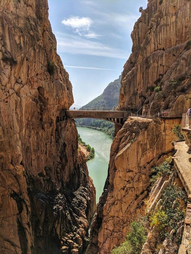 Un puente elevado conecta senderos construidos en las escarpadas paredes rocosas de un cañón profundo, con un río turquesa fluyendo abajo.