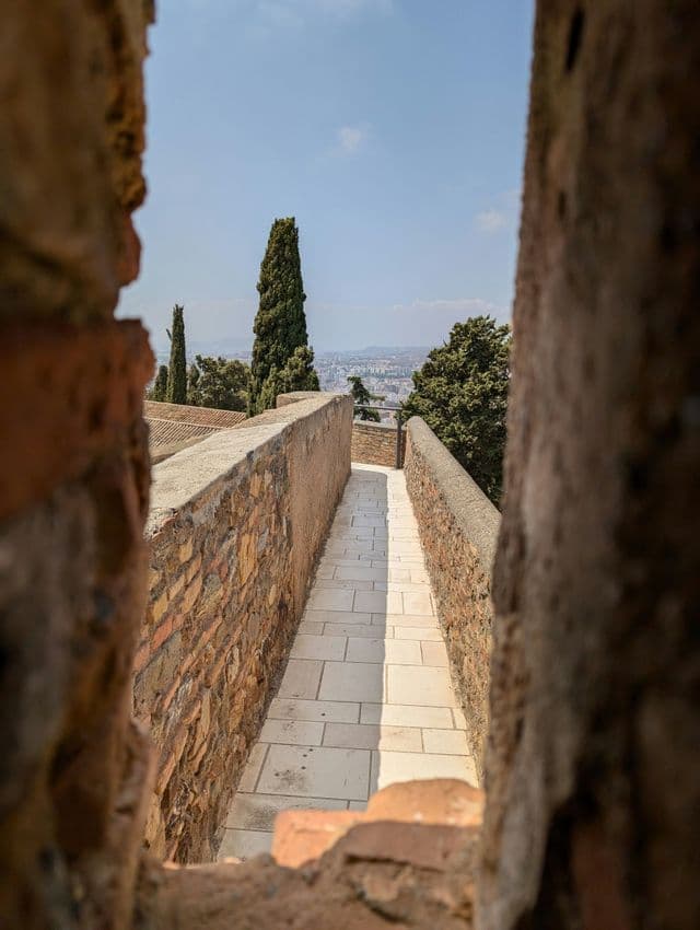 Un estrecho sendero pavimentado en una muralla de piedra, visto a través de una abertura, con vistas a un paisaje urbano con cipreses.