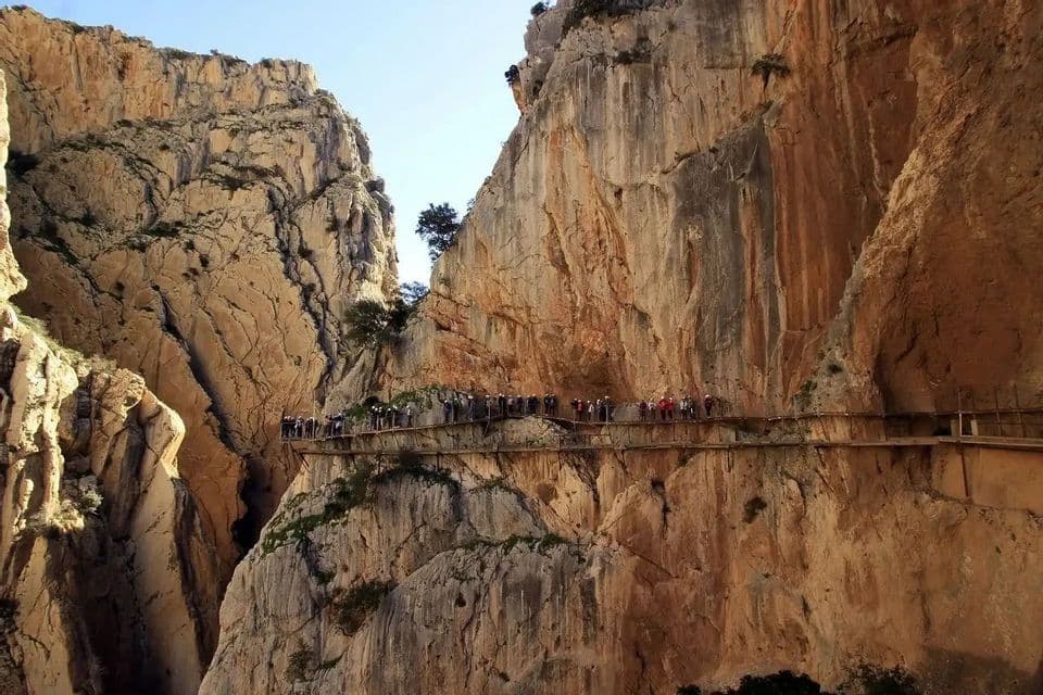 Eine WeRoad Gruppenreise wandert auf einem schmalen Holzsteg, der an einer steilen, felsigen Schluchtwand unter klarem Himmel befestigt ist.