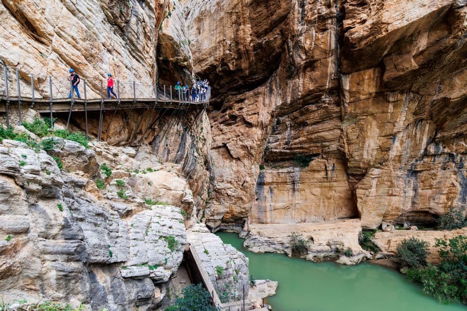 Eine WeRoad-Gruppe mit Helmen geht auf einem Holzsteg, der an einer Felswand in einer Schlucht befestigt ist, hoch über einem grünen Fluss.