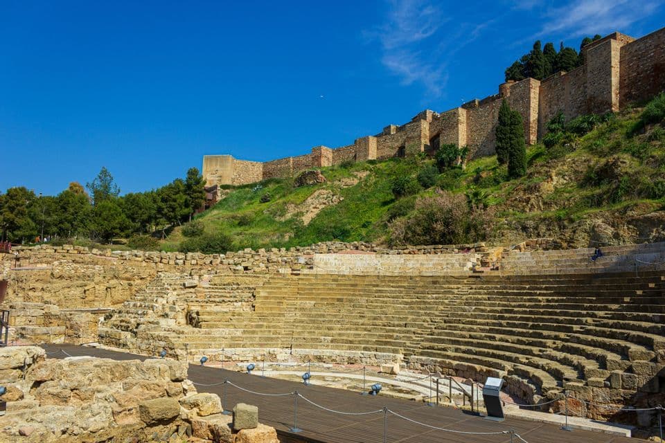 Die Ruinen eines steinernen Amphitheaters am Fuße eines grünen Hügels, mit einer großen Festungsmauer oben unter klarem blauem Himmel.