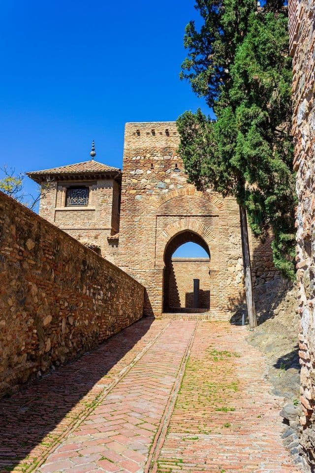 A brick path leads uphill towards an arched gateway in an ancient stone fortress under a clear blue sky.