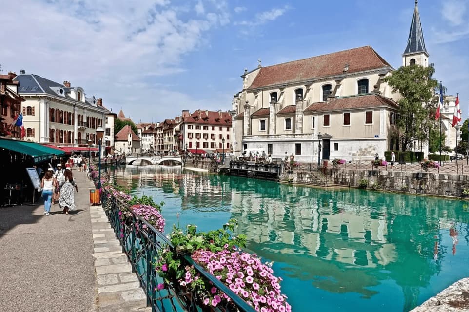 Persone camminano lungo un sentiero di pietra accanto a un canale turchese, con edifici storici e i loro riflessi visibili sotto un cielo azzurro.