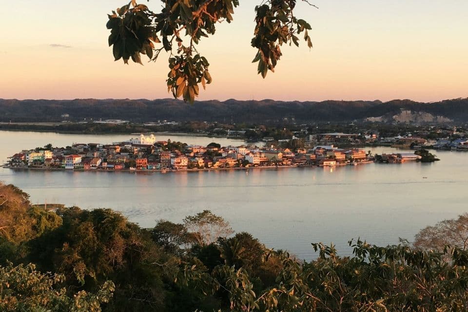 Blick aus der Vogelperspektive auf eine farbenfrohe Inselstadt an einem See, bei Sonnenuntergang von Baumblättern eingerahmt.