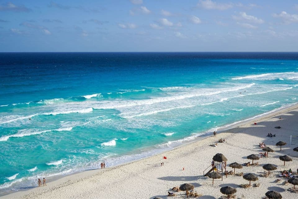 Eine Luftaufnahme eines weißen Sandstrandes mit Menschen, Strohschirmen und türkisfarbenen Meereswellen unter blauem Himmel.