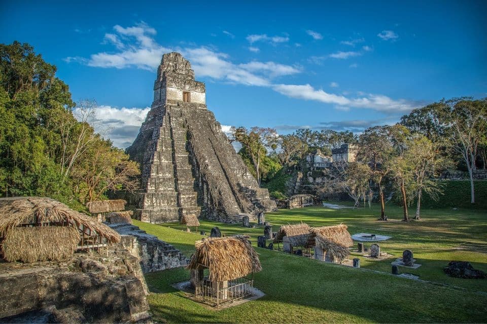 Eine große steinerne Stufenpyramide und antike Ruinen auf einer grasbewachsenen Fläche mit strohgedeckten Hütten, umgeben von Dschungel unter blauem Himmel.