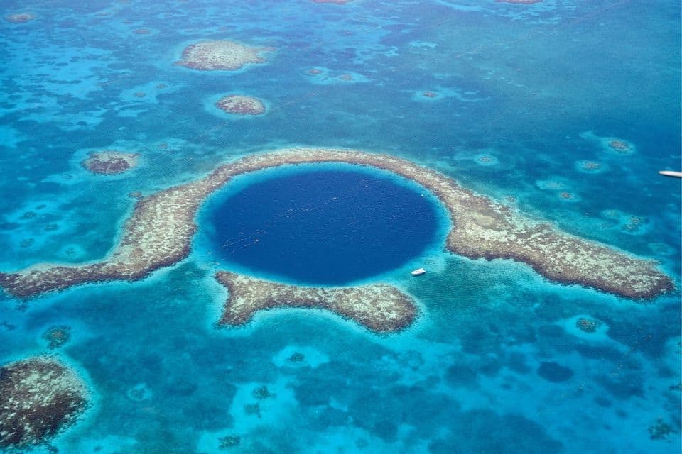 Eine Luftaufnahme eines großen, runden Blauen Lochs, umgeben von einem Korallenatoll in einem türkisfarbenen Ozean, mit einem kleinen Boot in der Nähe.