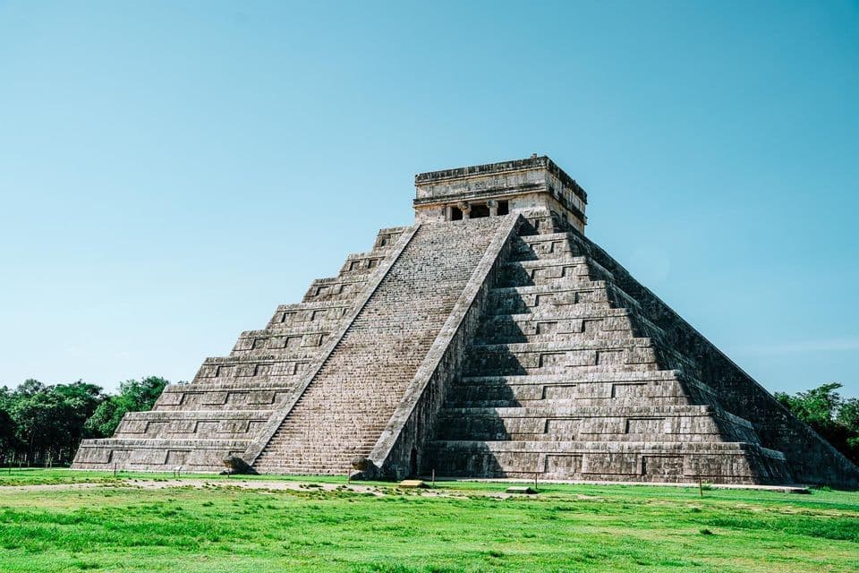 Eine große, alte Steinstufenpyramide mit einer steilen zentralen Treppe steht auf einem grünen Rasenfeld unter einem klaren blauen Himmel.