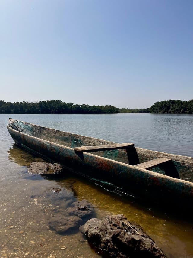 Ein verwittertes Holzkanu liegt im flachen Wasser eines Flusses, mit üppig grünen Bäumen am gegenüberliegenden Ufer unter klarem Himmel.